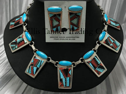 A Southwest Inlayed necklace and matching earrings set with sterling silver Inlayed with vibrant turquoise, coral, and Onyx geometric patterns. The necklace has multiple rectangular pendants, each with turquoise stones, and the earrings match the necklace design. Displayed on a black stand.