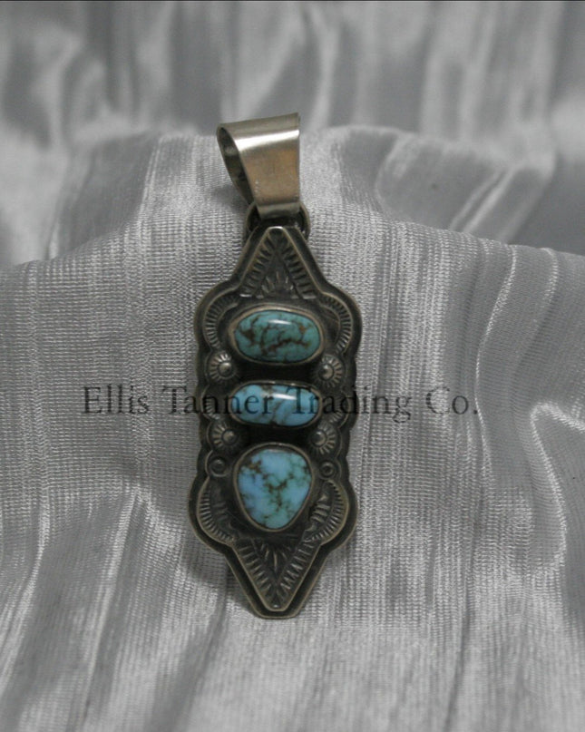 A hand stamped silver pendant with three turquoise stones arranged vertically, each set in a bezel. The pendant features intricate geometric and linear stamped patterns on the silver, It has a sturdy silver bail at the top and an elongated, curved shape.