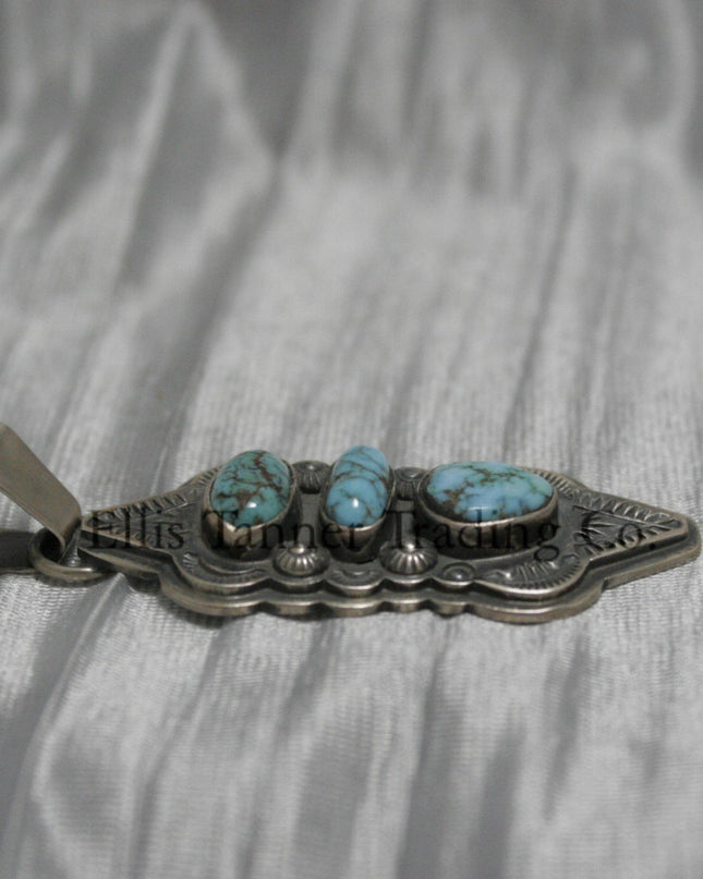 A hand stamped silver pendant with three turquoise stones arranged vertically, each set in a bezel. The pendant features intricate geometric and linear stamped patterns on the silver, It has a sturdy silver bail at the top and an elongated, curved shape.Side View 